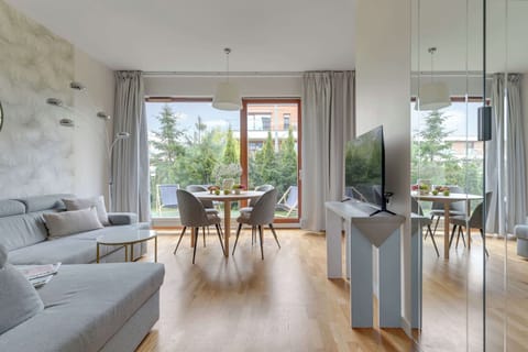 Nowoczesny salon z dużymi przeszkleniami i widokiem na zieleń tworzy jasną oraz komfortową przestrzeń do wypoczynku. Stylowe wnętrze łączy przytulną część wypoczynkową z funkcjonalnym aneksem kuchennym i jadalnią, zapewniając idealne warunki zarówno 