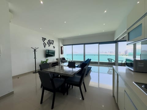 Dining area with dark wood table and chairs, adjacent to a glass balcony overlooking turquoise ocean waters.