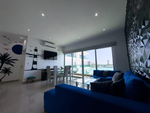 Living room features a deep blue sectional, geometric textured wall, glass dining table, and ocean views through sliding doors.