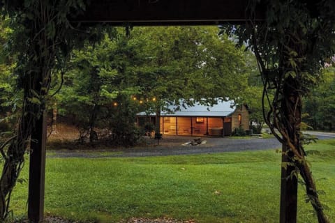 A charming cabin-style property nestled among mature trees and lush greenery, with warm lighting welcoming visitors at arrival.