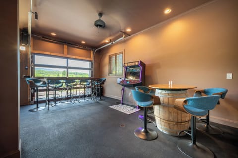 Features a classic arcade machine, a rustic barrel-style table with comfortable seating, and a high-top viewing area overlooking scenic outdoor views.