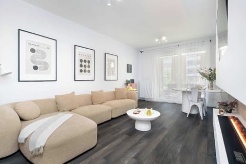 A bright living room features a large beige sectional sofa decorated with pillows, facing a minimalist white media console. Light wooden flooring and soft overhead lighting create an inviting, open atmosphere.