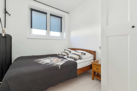 A compact bedroom features a single bed beneath a wide window that fills the space with natural light. Minimal furnishings and bright walls create a calm atmosphere.