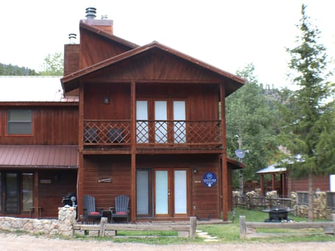 A mountain cabin property surrounded by forested hillsides, featuring a distinctive red-toned cedar exterior, brick chimney, lattice-accented balcony, and property signage, nestled in a scenic natural landscape.