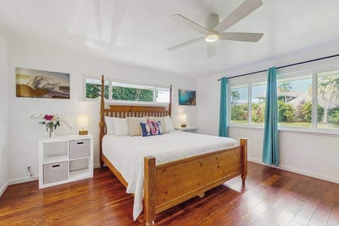 Coastal-style bedroom featuring a large wooden four-poster bed frame, warm hardwood floors, and vibrant teal curtains accenting wide windows that look out onto lush greenery.