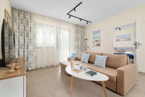 A cozy and bright living room features a comfortable beige sofa decorated with soft blue pillows and a sleek oval coffee table. Elegant floral curtains frame the window, while a flat-screen television sits on a white media console.
