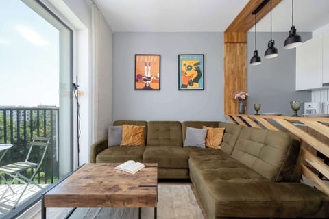 A stylish living room features a large, olive-green corner sofa decorated with warm orange accent pillows. A rustic wooden coffee table sits in front of it, while modern graphic art hangs on the grey wall next to a wooden slatted partition.