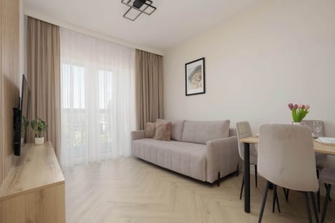 Picture 1: A bright living area features a comfortable beige sofa, a matching wooden dining set, and light streaming through large sheer curtains. The herringbone-patterned flooring adds an elegant and cohesive touch to the open space.