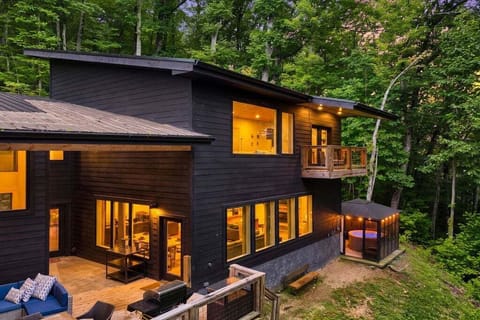 Evening exterior with private hot tub area, outdoor deck seating, and warm lighting surrounded by forest views.
