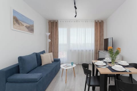 The open-plan living room is viewed from an angle that highlights both the blue sofa and the dining area. Bright textiles and light flooring enhance the sense of spaciousness.
