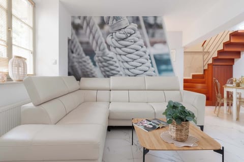 A bright living room features a large cream-colored sofa, a coffee table, and a staircase leading to the upper floor. Modern lighting and light finishes create an elegant atmosphere.