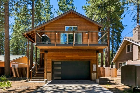 Two-story wood-clad house with black metal garage door, balcony railing, and exterior staircase surrounded by tall pine trees.