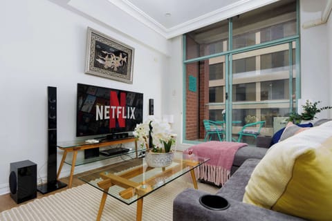 Bright, inviting living room with a comfortable sofa, smart TV for streaming, and a glass coffee table. Floor-to-ceiling windows open to a private balcony with seating, filling the space with natural light and a relaxed feel. 