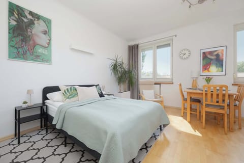 A bright studio apartment combines a comfortable bed, dining table, and kitchenette in one open space. Large windows fill the room with natural light.