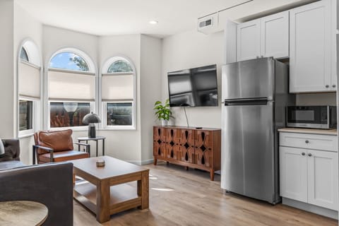 Open living area with seating, coffee table, TV, arched windows and an adjoining white kitchenette.