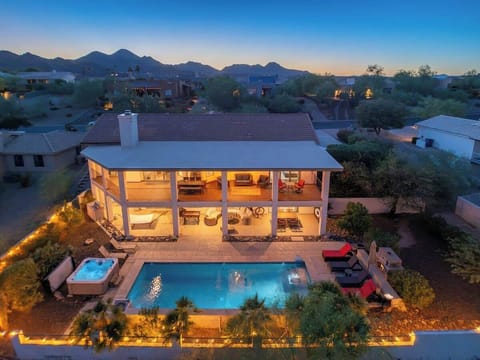 Stunning desert retreat showcases a modern two-story villa with an illuminated pool and hot tub, nestled in a tranquil neighborhood with dramatic mountain views at twilight.