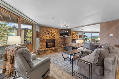 Unwind in your spacious mountain retreat with a stunning stone fireplace, cozy seating, and panoramic views framing the nearby peaks through floor-to-ceiling windows.