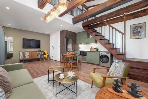 A spacious open-concept living area features a green sofa, an armchair, and round coffee tables arranged on a patterned rug. Wooden stairs lead up to a loft level, while a compact kitchen and dining area occupy the background.