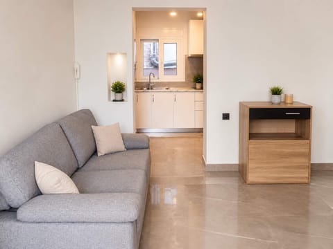 A clean living area with a comfortable grey sofa, polished floors, and the visible kitchen in the background. The space feels modern and simple, ideal for a relaxed stay