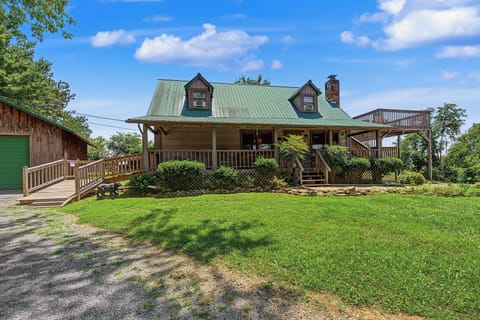 Charming mountain cabin with a green metal roof, wraparound deck, and lush lawn set against mature trees and blue skies.