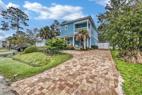 A charming coastal home with bright blue siding welcomes guests, featuring a paver driveway, lush tropical landscaping, and an inviting second-story balcony perfect for relaxing in the sunny Florida weather.
