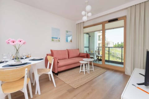 A bright living room features a coral-pink sofa on a woven rug next to a white dining set. Large sliding glass doors lead out onto a sunny balcony.