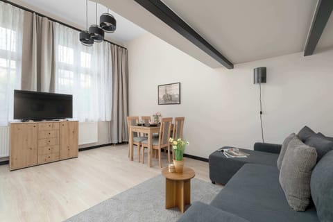 A spacious living room features a dark grey corner sofa paired with a small wooden coffee table. A wooden dining table sits in the background near a large window, while a TV unit is positioned opposite the seating area.