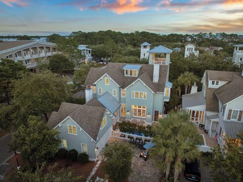 Aerial view of a charming coastal vacation home nestled in a serene tree-lined neighborhood, with warm interior lighting glowing as dusk settles over the surrounding landscape.