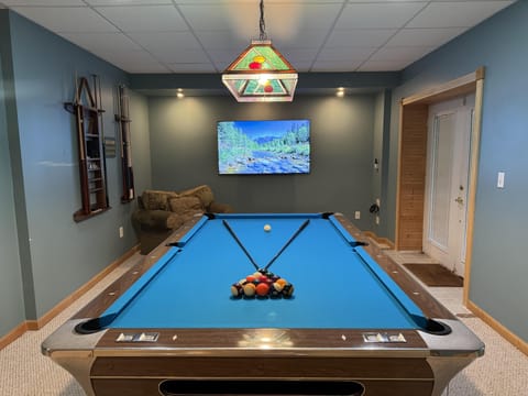 Game room with blue felt pool table, wall-mounted flat-screen TV, plush armchair, and stained-glass pendant light.