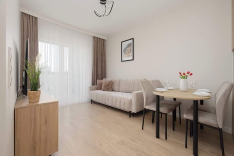 A bright living room features a beige sofa and a small round wooden dining table. Glass doors at the back lead out onto a sunny balcony.