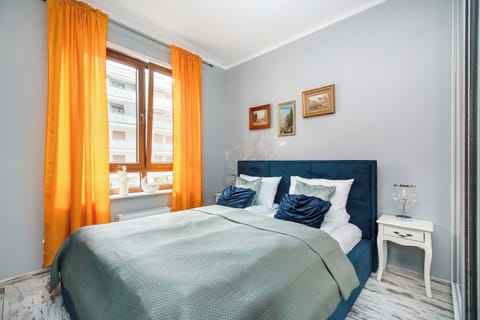A cozy bedroom features a double bed with a dark blue tufted headboard and a light teal bedspread. Bright orange curtains frame a large window, adding a vibrant splash of color to the otherwise neutral gray walls.