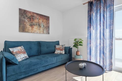 A vibrant teal-blue sofa sits on light wood flooring next to a black circular coffee table. Patterned curtains frame the balcony window in the corner.