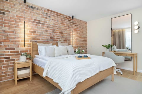 A cozy bedroom features a double bed with crisp white linens and a wooden frame set against a warm brick accent wall. A sleek vanity desk with a circular mirror stands to the right, adding functionality to the modern space.