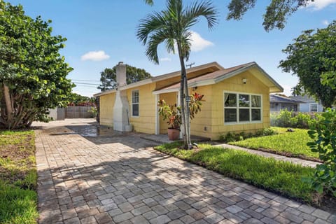 Charming yellow cottage exterior of The Banana Cabana, featuring a paver driveway, lush palm trees, and inviting curb appeal in West Palm Beach, FL