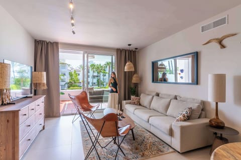 The bright living room with a dining area is decorated in natural tones and a Mediterranean style. It features a sofa, two armchairs, a Smart TV, and a dining table with chairs. Large windows lead to a terrace overlooking the pool.