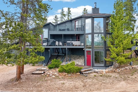 Modern lakeside retreat framed by towering pines, featuring striking architecture with expansive windows, multiple balconies, and a distinctive glass entrance tower with red accents.