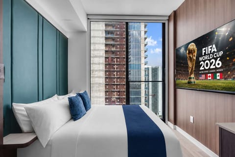 Bedroom with a plush queen bed, upholstered headboard, and floor-to-ceiling windows framing the Miami skyline. Smart TV mounted opposite — catch every World Cup 2026 match from bed.