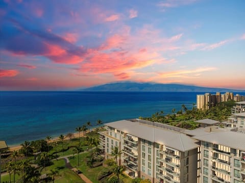 Stunning beachfront resort with vibrant sunset skies over tropical waters and distant mountain silhouette.