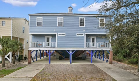 A charming two-story duplex on stilts with a distinctive blue and white exterior, featuring dual porches and covered parking, nestled in a residential coastal neighborhood.