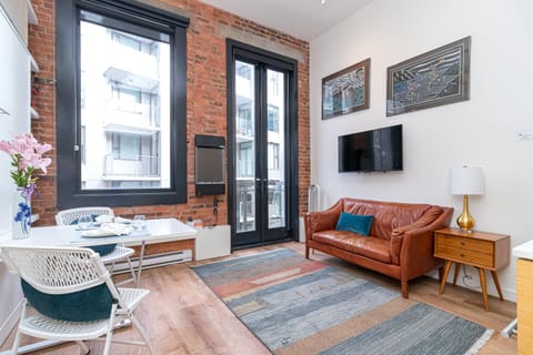 Exposed brick walls frame a leather sofa and mid-century nightstand beneath a wall-mounted TV in the living area.
