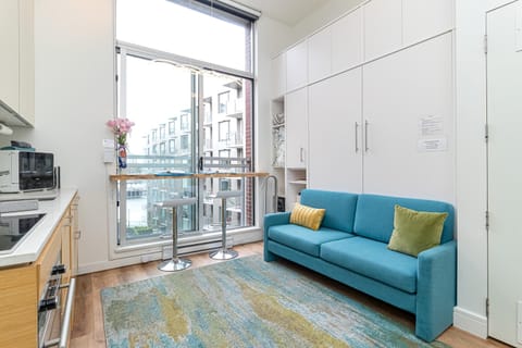 A blue sofa with yellow and green pillows faces a tall window and breakfast bar with two metal stools.