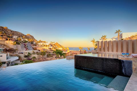 An infinity pool overlooking a hillside coastal community during golden hour. The property sits among terraced homes carved into the rocky landscape, with ocean views stretching to the horizon.