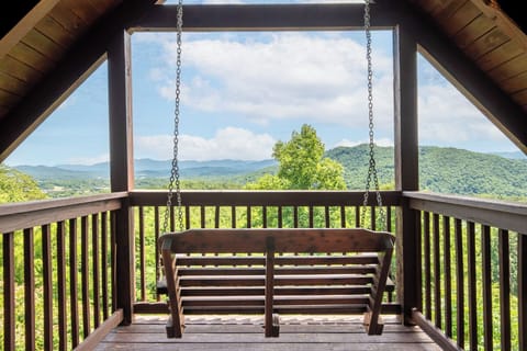A covered porch swing framed by rolling green mountains under a bright blue sky, offering scenic mountain views and a peaceful spot to relax.