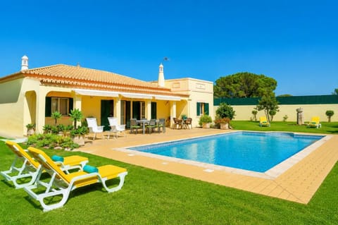 A ground-level view of a single-story villa featuring a rectangular swimming pool, a spacious tiled patio with outdoor seating, and a manicured green lawn with yellow loungers under a bright blue sky.