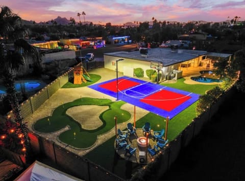 As the lights come on, the whole backyard turns into one big invitation — court, green, fire pit lounge, and string lights overhead.