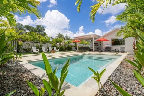 Private backyard oasis with a sparkling pool, lounge seating, and shaded dining under red umbrellas. Surrounded by lush tropical landscaping for a resort feel right at home.