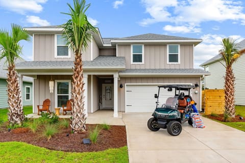 A welcoming two-story coastal-style home with palm-framed entrance, covered front porch seating, and convenient golf cart parking for easy neighborhood exploring.