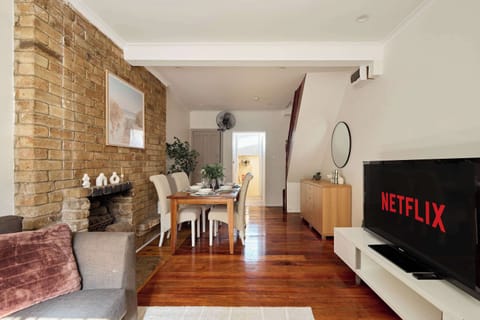 Relax in the cozy open-plan living and dining area, featuring exposed brickwork, warm timber flooring, a smart TV with streaming options, and an inviting dining setup.