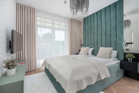 A bedroom features a double bed with a teal velvet channel-tufted headboard. Sheer white curtains and soft pink drapes frame the large balcony window.