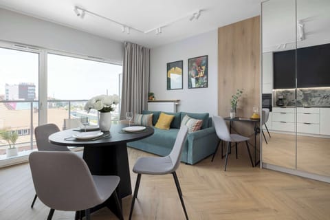 A modern living area features a round black dining table with grey chairs next to a teal sofa. Large windows and mirrored wardrobe doors brighten the open space.